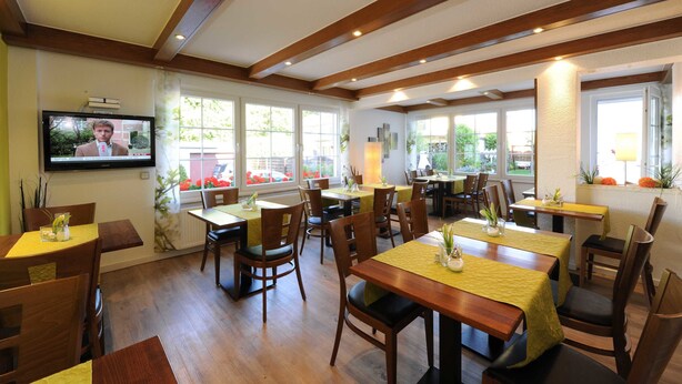 A dining area with wooden tables and chairs, large windows, and a television on the wall. - Hotel Garni Schilling (Duisburg)
