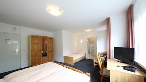 A hotel room with a bed, a desk with a TV, a wooden wardrobe, and a small seating area. - Hotel Garni Schilling (Duisburg)