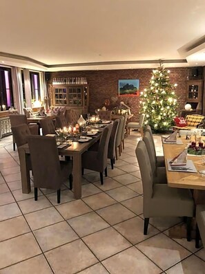 A dining area with a long table set for a meal, surrounded by chairs. A Christmas tree is visible in the background.