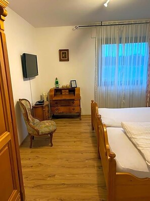 A bedroom with a wooden bed, a chair, a dresser, a flat-screen TV, and a window with curtains.
