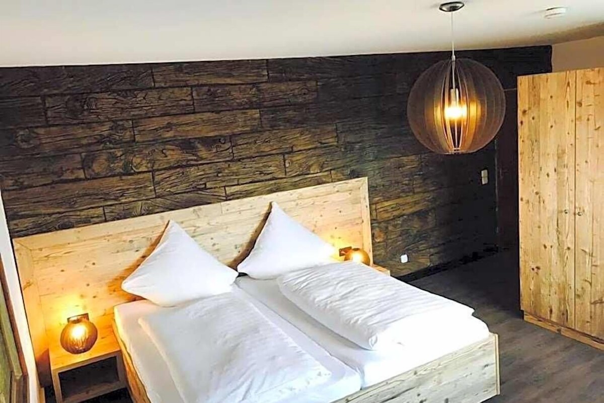 Quarto com cabeceira de madeira, cama com roupa de cama branca, luminária pendente e guarda-roupa de madeira.