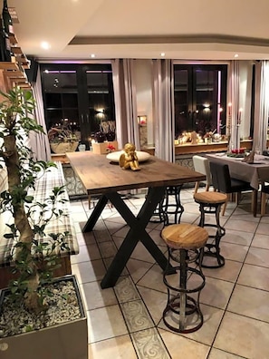 A dining area with a wooden table and bar stools, surrounded by potted plants and a view of the night sky.