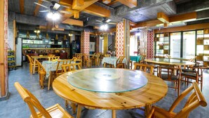 Ein Restaurant mit Holztischen und -stühlen, einer Küchenzeile und gemusterten Vorhängen.
