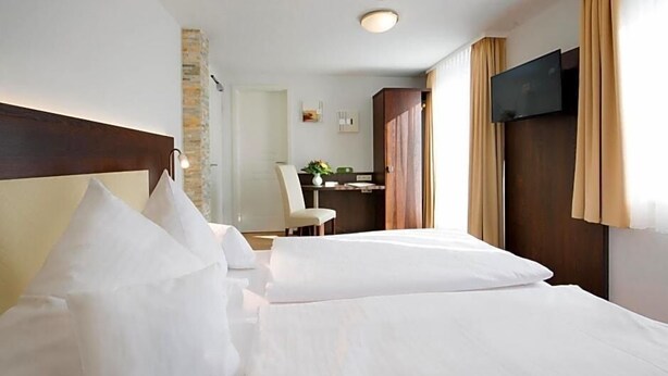 A hotel room with a bed, a desk, a chair, a TV, and a stone wall. - Birkenfelder Hof (Birkenfeld)