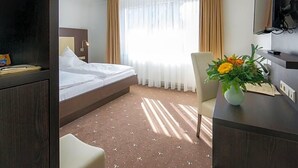A hotel room with a bed, a desk with a vase of flowers, a television, and a window with curtains. - Birkenfelder Hof (Birkenfeld)