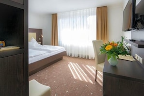 A hotel room with a bed, a desk with a vase of flowers, a television, and a window with curtains. - Birkenfelder Hof (Birkenfeld)