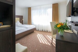 Una habitación de hotel con cama, escritorio con un florero, televisor y ventana con cortinas.