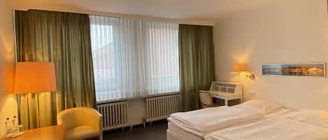 A hotel room with a bed, a chair, a desk, a lamp, a radiator, and a window with curtains.