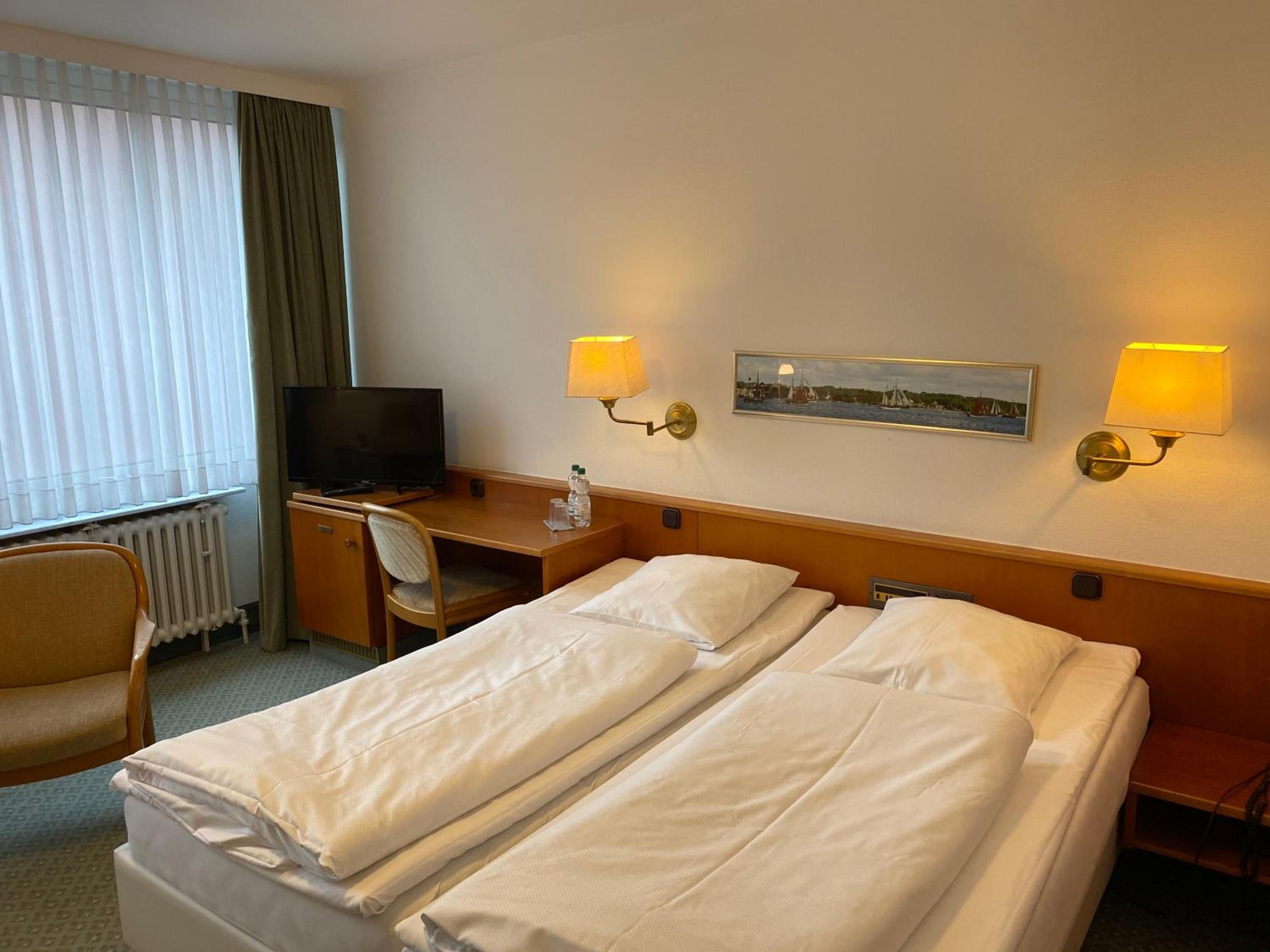 A hotel room with a bed, a desk, a chair, a television, and two wall-mounted lamps.