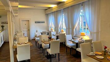 Un comedor de hotel con varias mesas y sillas, ventanas amplias con cortinas y suelo de ladrillo.
