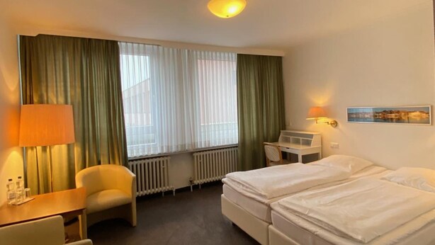 A hotel room with a bed, a chair, a desk, a lamp, a radiator, and a window with curtains.