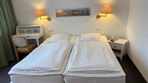 A hotel room with two single beds, a desk, a chair, a nightstand with a phone, and two wall-mounted lamps.