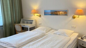 A hotel room with a double bed, two bedside lamps, a desk with a chair, a radiator, and a framed picture on the wall.