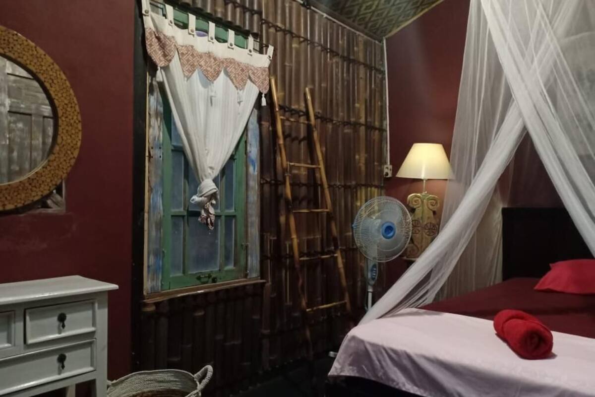 Een kamer met een bed, nachtkastje, lamp, ventilator en een raam met gordijnen.