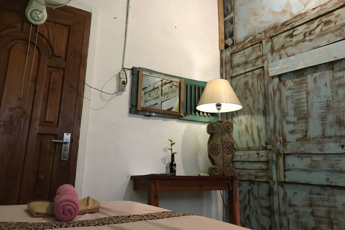 Een kamer met een bed, een houten deur, een ventilator aan de muur, een tafel met een lamp en een spiegel.