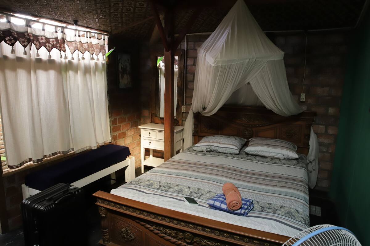 Een slaapkamer met een bed, nachtkastje, koffer, ventilator en een raam met gordijnen.