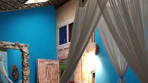 Une chambre avec un mur bleu, un lit recouvert d’une couverture à carreaux, une armoire en bois, un ventilateur et un rideau.