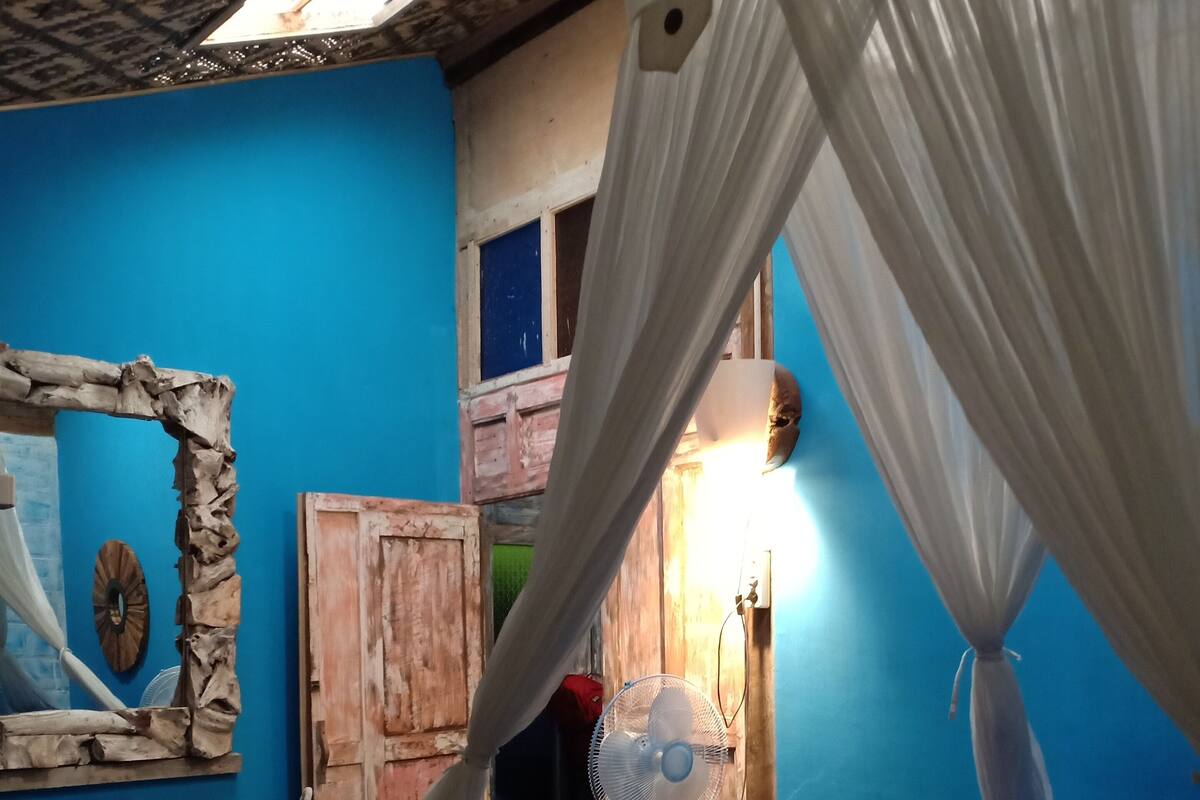 Een slaapkamer met een blauwe wand, een bed met een geruit dekbed, een houten garderobe, een ventilator en een gordijn.