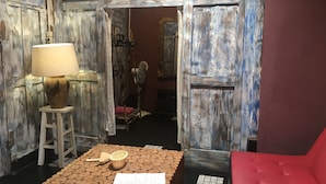 Une pièce avec une porte en bois rustique, un banc rembourré de coussins rouges, un tabouret en bois avec une lampe et une petite table sur laquelle se trouvent un panier et une feuille de papier.