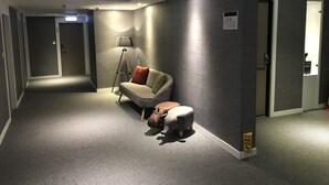 A modern hotel corridor with a grey carpet, a sofa with cushions, a floor lamp, and a baby walker.
