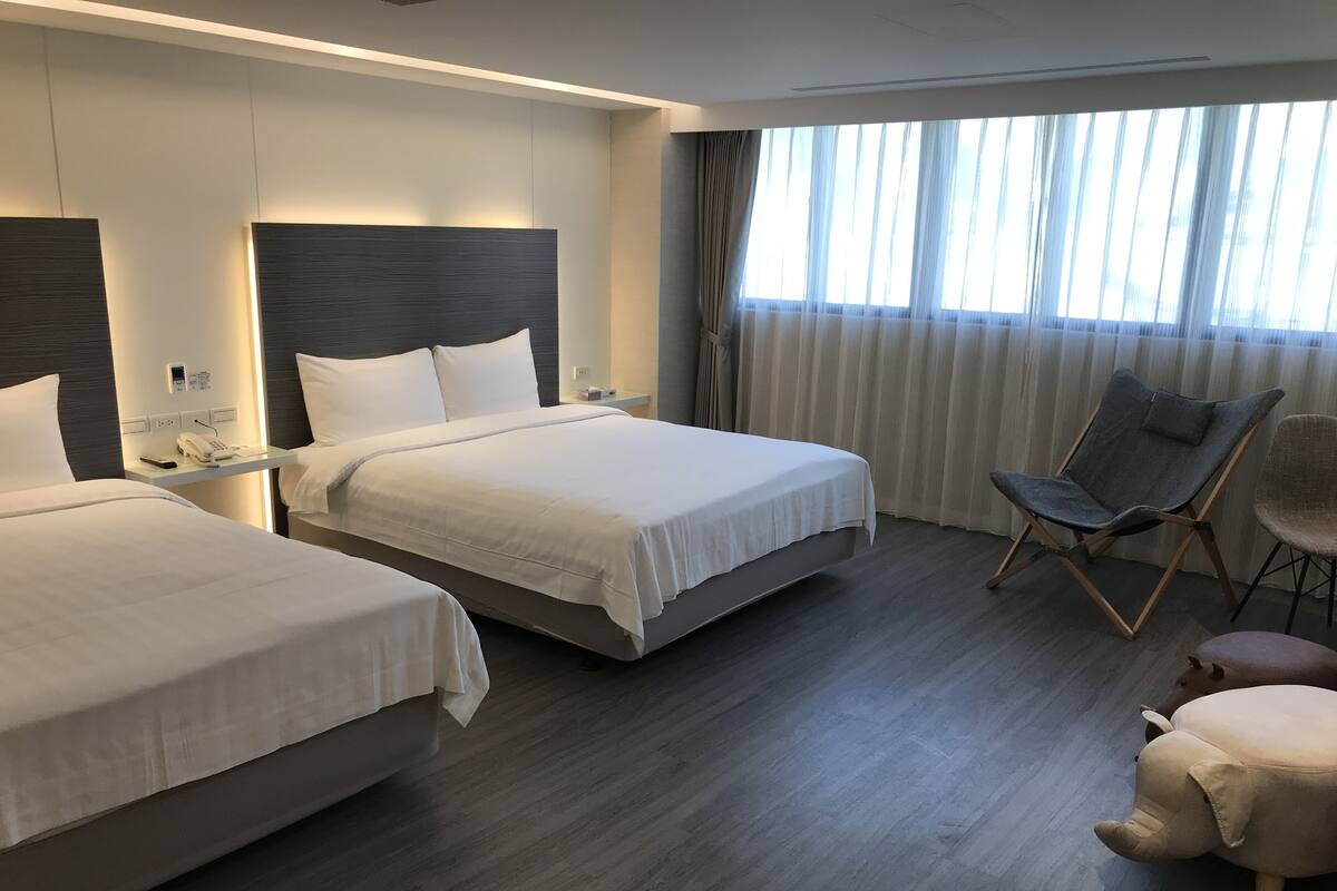 A hotel room with two beds, a desk with a phone, a chair, and a large window with curtains.