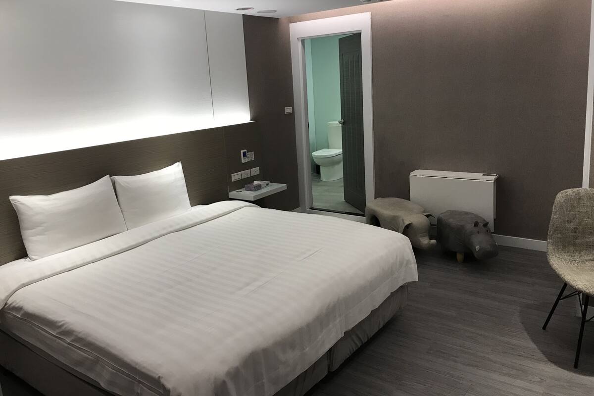 A modern hotel room with a large bed, bedside table, chair, and a small stool.