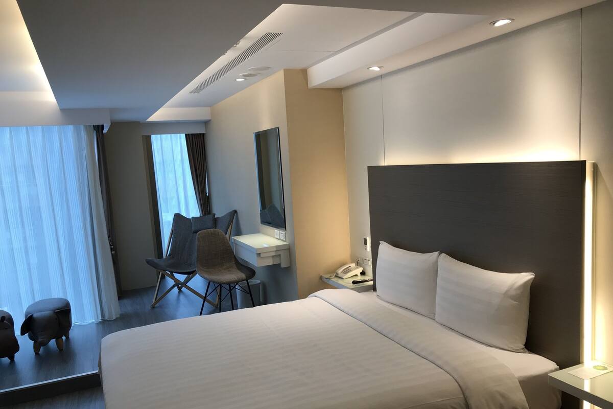 A modern hotel room with a large bed, a desk with a phone, a chair, and a mirror.