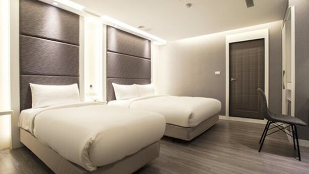 A hotel room with two beds, a dark headboard, a chair, and a door.
