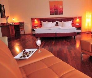 Une chambre d’hôtel avec un grand lit, deux lampes de chevet, un plancher en bois et une petite table sur laquelle se trouvent un verre d’eau et des chocolats.