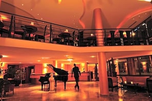 A spacious lobby with a grand piano, a reception desk, and a balcony with seating. - Hotel Santo (Karlsruhe)
