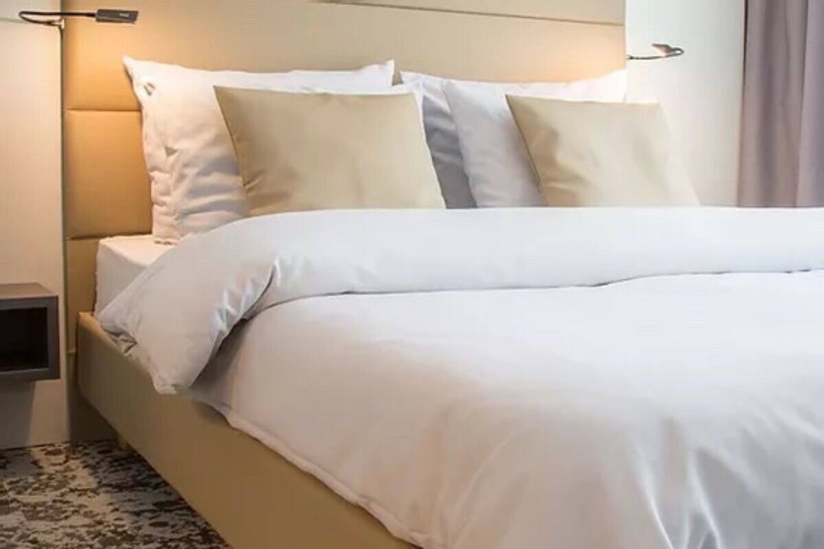 A neatly made bed with white linens and a headboard, a bedside table, and a lamp.