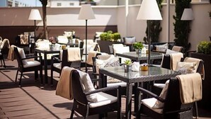 An outdoor dining area with wicker furniture, white cushions, and hanging lamps. - Hotel Santo (Karlsruhe)