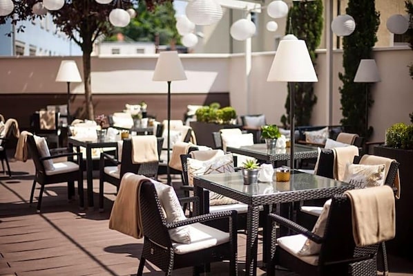 An outdoor dining area with wicker furniture, white cushions, and hanging lamps. - Hotel Santo (Karlsruhe)