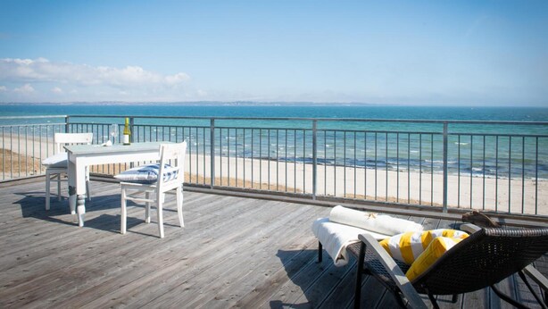 A wooden deck with a table and chairs, overlooking a beach and the sea. - Strandhotel Lodge am Meer (Gluecksburg)