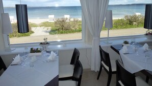 A dining area with tables set for two, large windows with white curtains, and a view of the beach and sea. - Strandhotel Lodge am Meer (Gluecksburg)