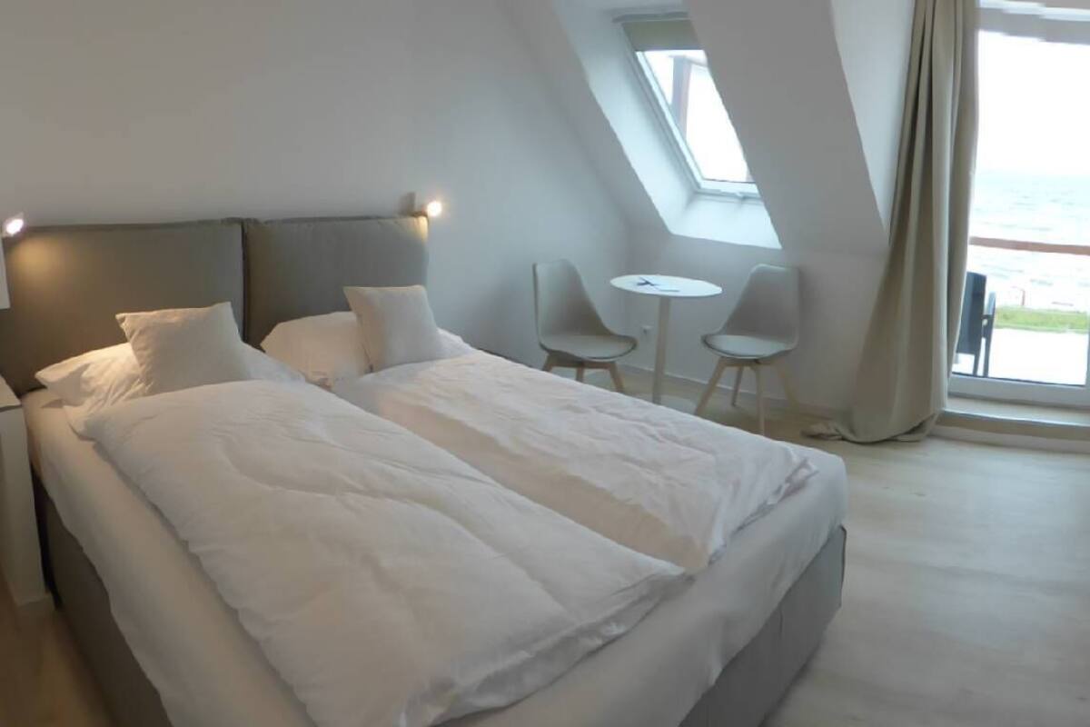 Une chambre moderne avec un grand lit, une petite table et des chaises, et une vue sur la mer par une grande fenêtre.