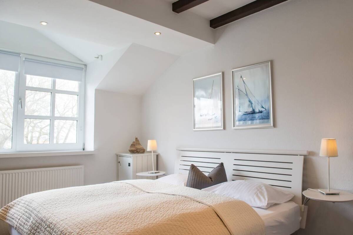Une chambre à coucher avec un grand lit, une fenêtre, un radiateur, une table de chevet, une lampe et deux tableaux encadrés accrochés au mur.
