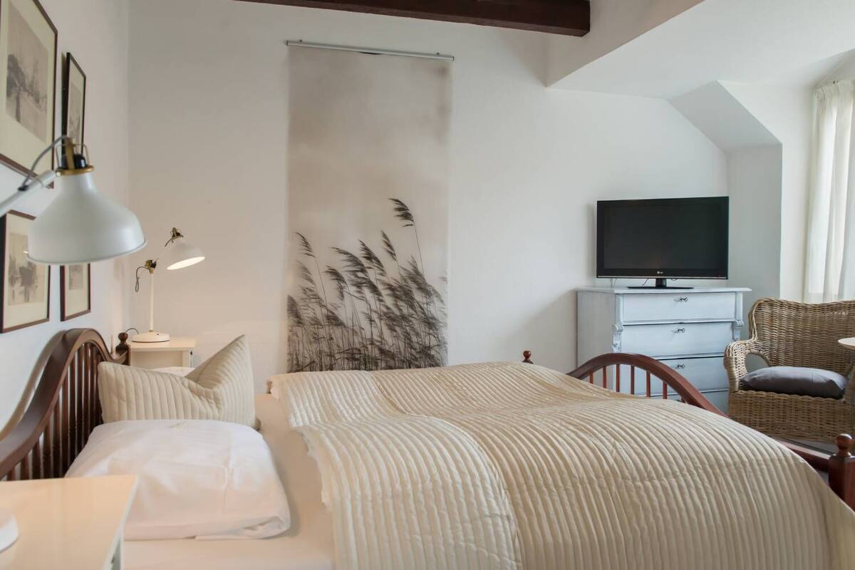 Une chambre à coucher comprenant un lit, des lampes de chevet, un téléviseur, une commode et un fauteuil en osier.