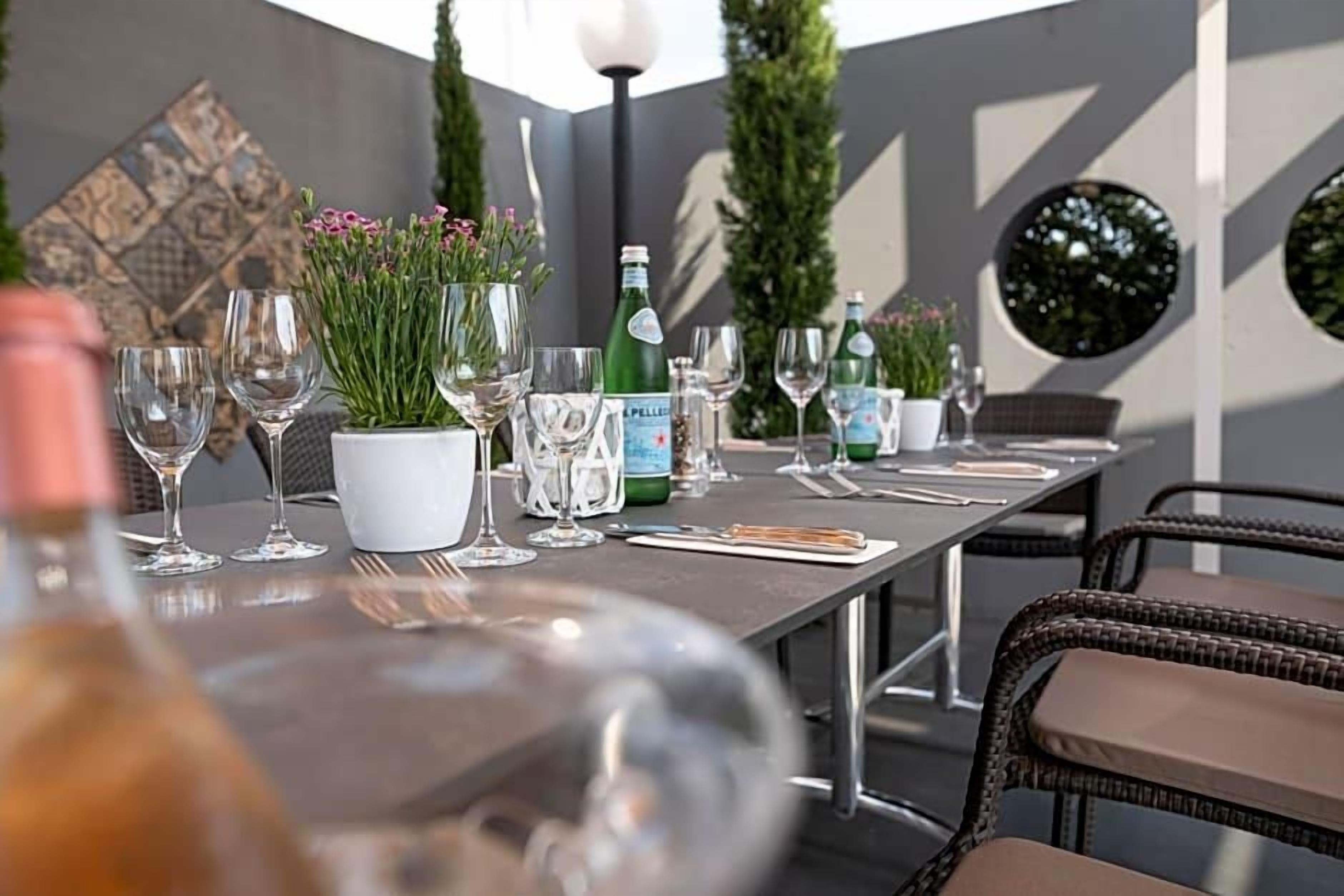 An outdoor dining area with tables set for two, featuring clear glassware, potted plants, and bottles.
