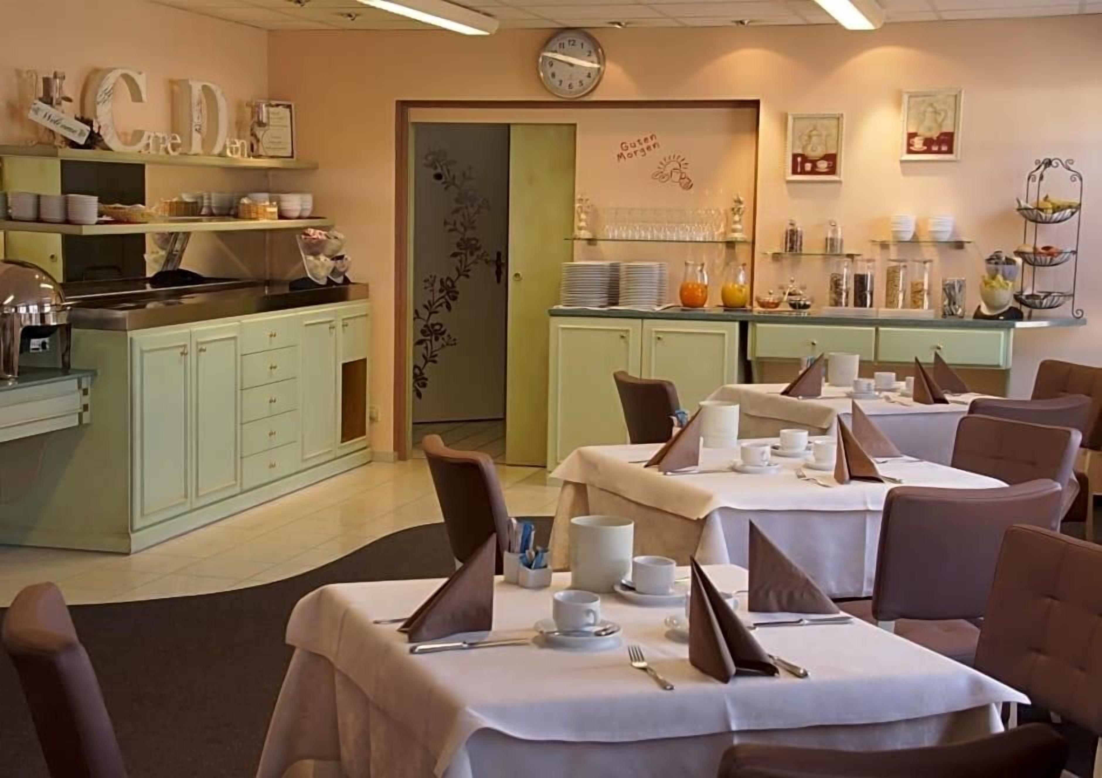 A dining area with tables set for service, a kitchen area with a clock, and a wall with decorative elements.
