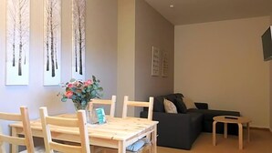 Una sala de estar con una mesa de comedor, sillas, un sofá, una mesita auxiliar y un televisor montado en la pared.
