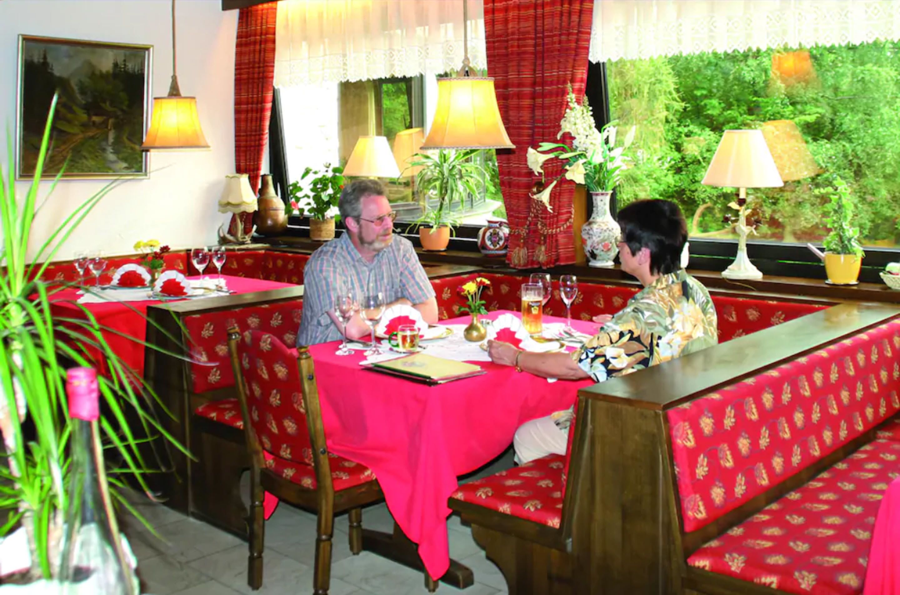 A restaurant with red upholstered booths, a table set with wine glasses and a menu, and potted plants.