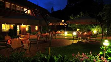 An outdoor patio area with tables and chairs under umbrellas, surrounded by greenery.