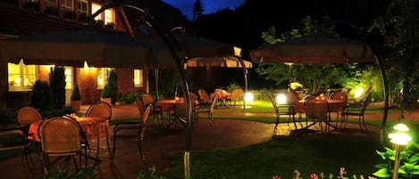 Un área de patio exterior con mesas y sillas bajo sombrillas, rodeada de follaje.