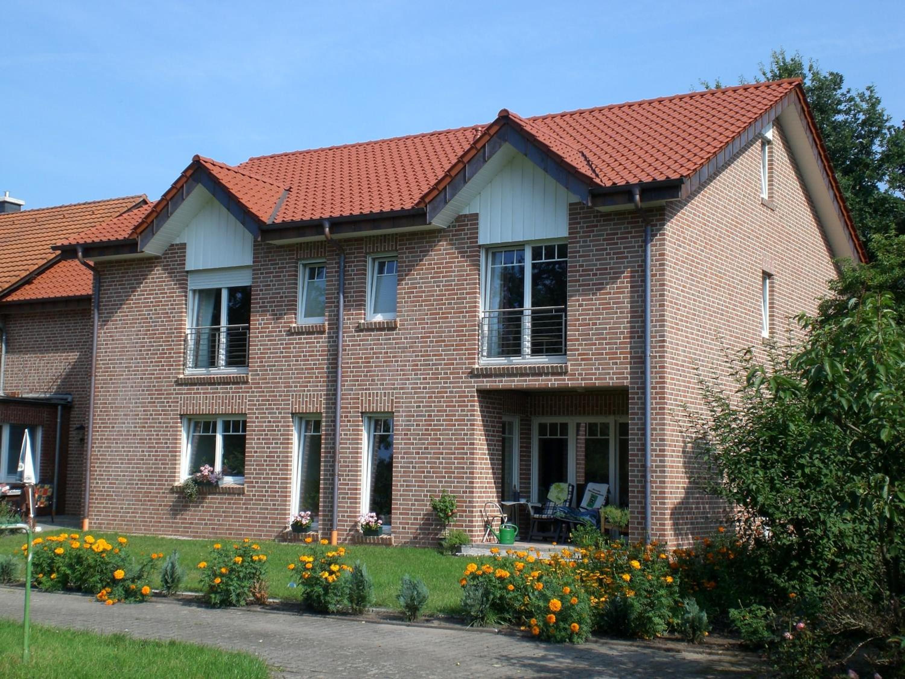 A two-story brick house with a red-tiled roof, a front porch with a bench, and a garden with marigolds.