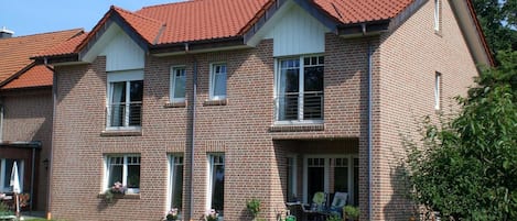A two-story brick house with a red-tiled roof, a front porch with a bench, and a garden with marigolds.