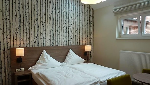 Un lit double avec deux oreillers, une table de chevet avec une lampe, une table ronde avec une chaise, et une fenêtre donnant sur des immeubles.