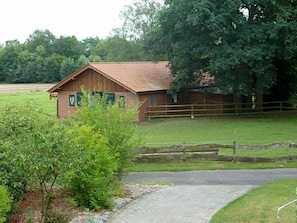 A rustic wooden building with a sloped roof, surrounded by a fenced pasture and a paved path leading to it.