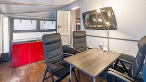 Ein kleines Zimmer mit einem Tisch, vier Stühlen, einem roten Schrank, einem Fenster und einem an der Wand befestigten Fernseher.
