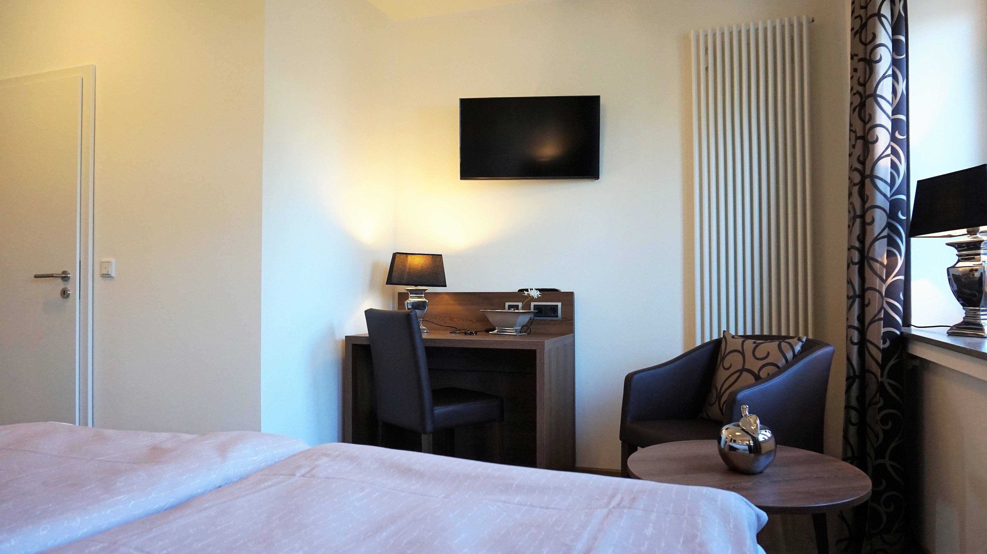 Habitación de hotel con una cama, un escritorio con lámpara, una silla, una mesita y un televisor en la pared.